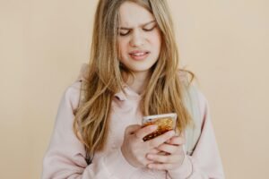 Teen girl with blonde hair in pink hoodie using smartphone, expressing curiosity indoors.